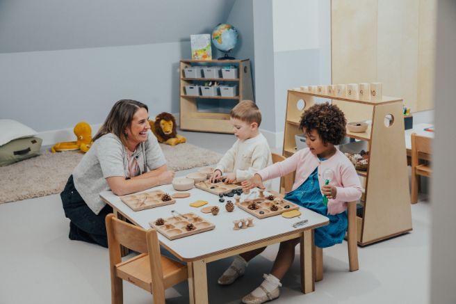 Two children playing with toys on a table and educator sitting next to them and smiling