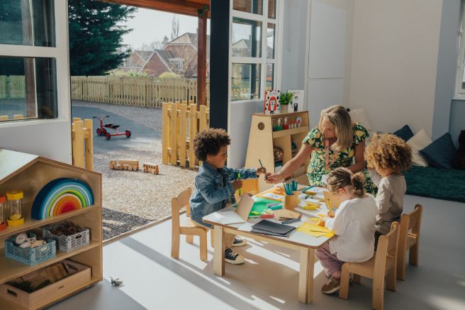 Children sitting at a desk in a nursery and drawing with their educator