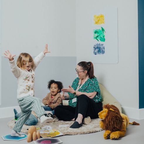 An educator sitting on the floor in a nursery next to a child while another child jumps around happily