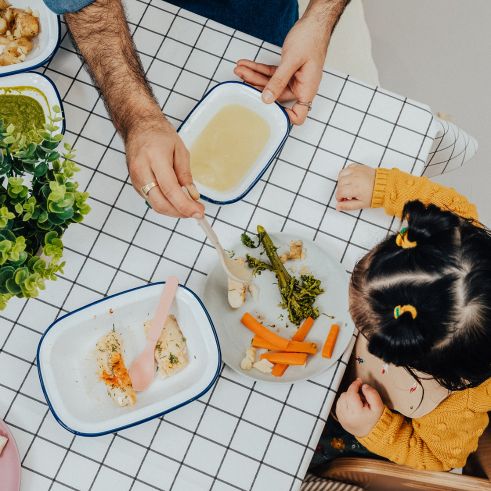 A toddler eating at a table in a nursery, an educator is feeding her