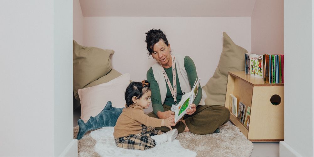 An educator reading to a toddler while sitting on the floor in a nursery playroom