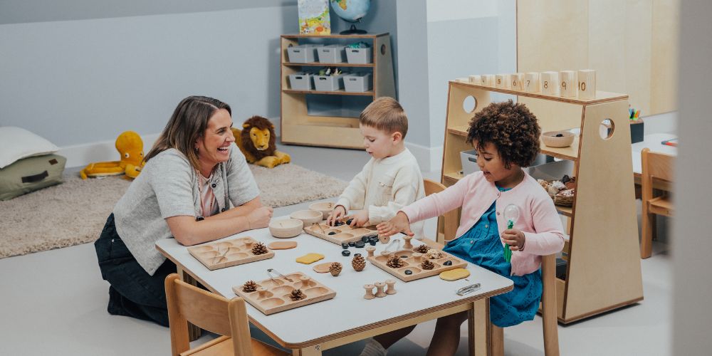 Two children playing with toys on a table and educator sitting next to them and smiling