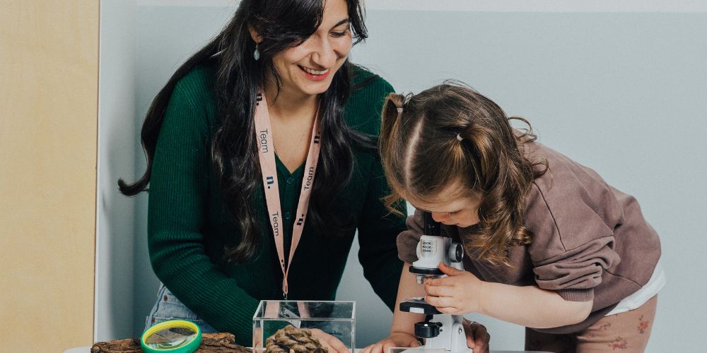 An educator sitting next to a child while the child looks through a microscope