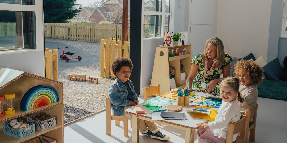 Children sitting at a desk in a nursery and drawing with their educator