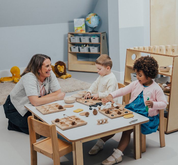 Two children playing with toys on a table and educator sitting next to them and smiling