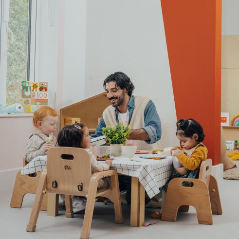 A nursery educator sitting around a toddler table with toddlers feeding them