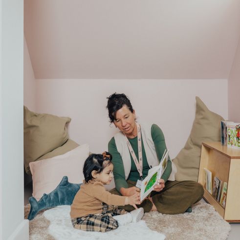 An educator reading to a toddler while sitting on the floor in a nursery playroom