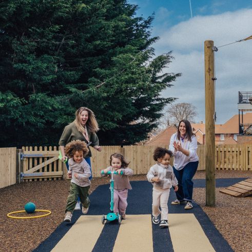 Two educators cheer as three young children joyfully run and ride scooters across a playground