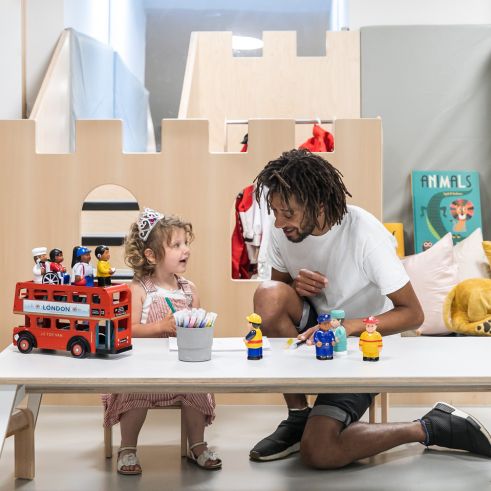 An educator kneels beside a young girl wearing a tiara as they play together at a small table with toy figures and a red London bus