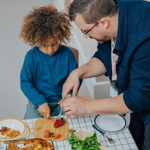 Male early years educator helping a young child slice strawberries during a cooking activity at N