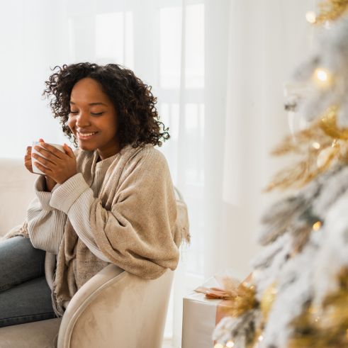 Early years educator relaxing at home with a warm drink beside a softly decorated winter tree.