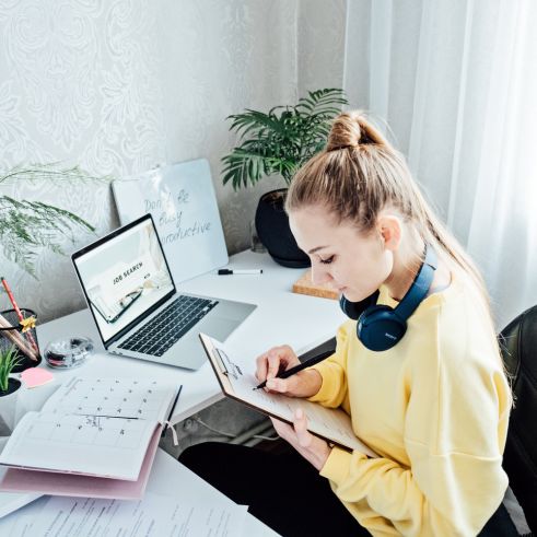 Young woman sitting at a desk and updating her CV, surrounded by notebooks and a laptop.