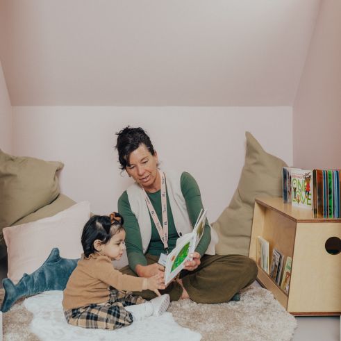 A female educator sitting on a rug in a cozy reading nook, reading a picture book to a child.