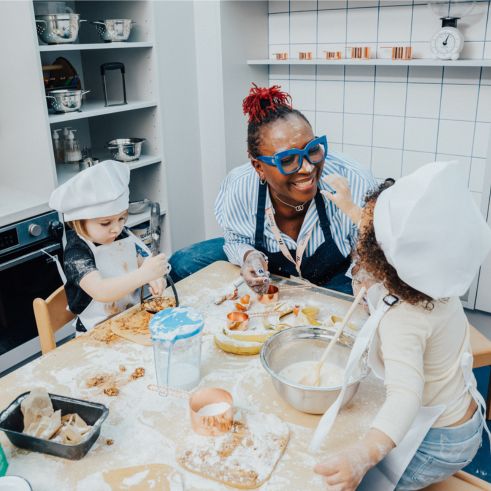 An early years educator smiling and baking with two young children at a small table in a nursery kitchen.